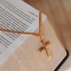 Gold-tone cross necklace elegantly draped over the edge of an open Bible, highlighting the cross pendant against the gilded pages in a warm, faith-centered setting that symbolizes devotion and trust in God’s Word.