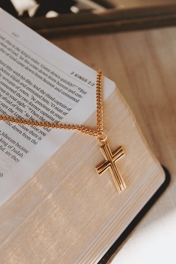 Gold-tone cross necklace elegantly draped over the edge of an open Bible, highlighting the cross pendant against the gilded pages in a warm, faith-centered setting that symbolizes devotion and trust in God’s Word.