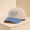 Beige and light blue Christian baseball cap embroidered with the phrase “Please Ask Me About My God,” displayed on a minimalist light wood pedestal against a neutral beige background.