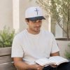 Man wearing a white and black baseball cap with an embroidered cross while reading his Bible on an outdoor bench in a peaceful setting.