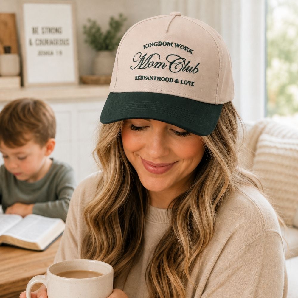 Mother wearing a “Mom Club – Kingdom Work, Servanthood & Love” Christian baseball cap while enjoying coffee at home as her child reads a Bible in the background.