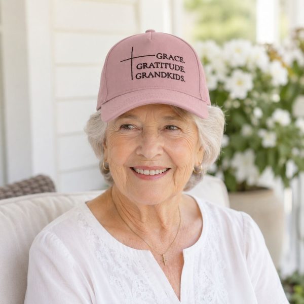 Smiling elderly woman wearing a pink Christian hat embroidered with “Grace, Gratitude, Grandkids” while relaxing on a bright porch.