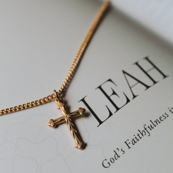 Beautiful gold-tone cross necklace resting on a page featuring the word “LEAH” and the phrase “God’s Faithfulness,” creating a soft and elegant devotional setting. The delicate cross design and warm gold finish symbolize faith, hope, and spiritual encouragement, making it a meaningful gift or everyday expression of Christian belief.