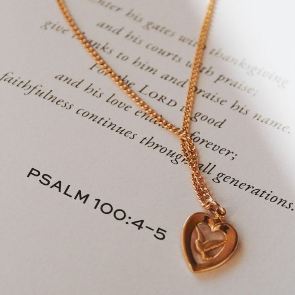 Gold-tone heart pendant necklace featuring a dove design, gracefully displayed on an open Bible page beside Psalm 100:4–5, creating a warm and meaningful Christian devotional setting.