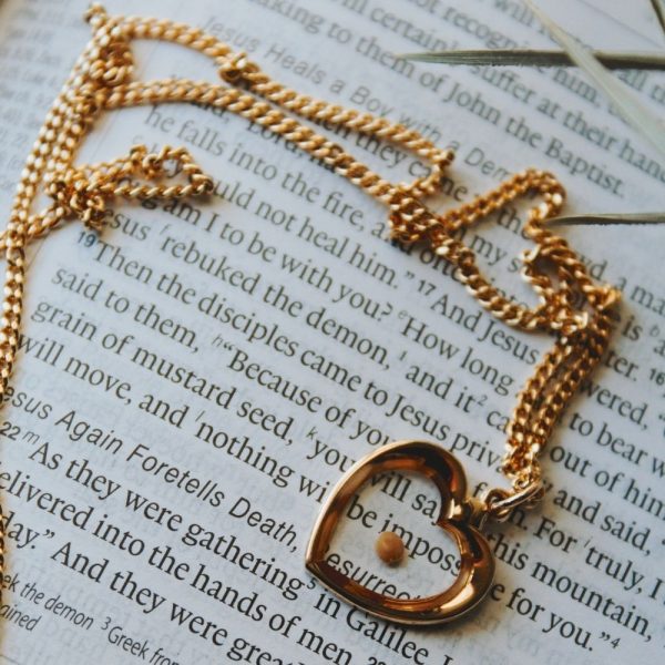 Gold heart pendant necklace resting on an open Bible, Christian faith jewelry for women, elegant heart charm necklace with Scripture background, inspirational Christian gift, devotional jewelry, faith-based necklace, religious gift for women, rose gold Christian necklace, meaningful everyday faith accessory.