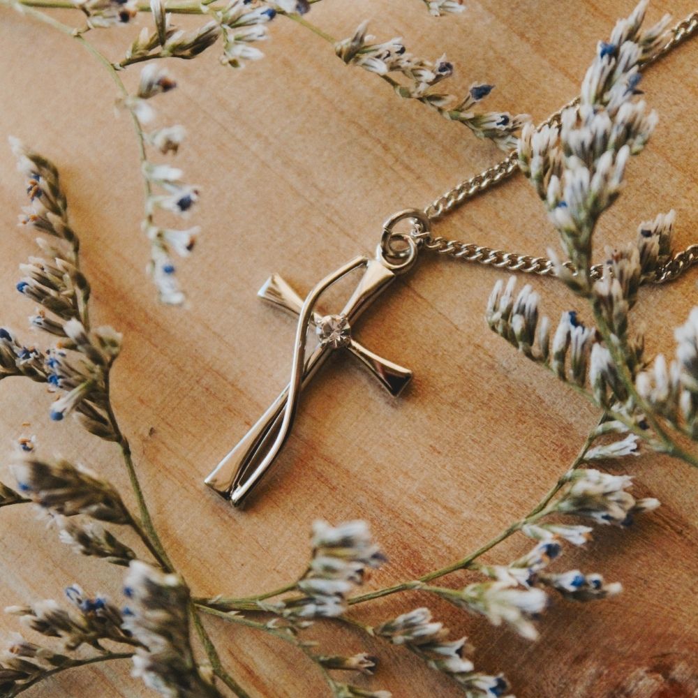 Twisted cross pendant necklace with crystal accent resting on a natural wood surface surrounded by delicate wildflowers, Christian faith jewelry, elegant cross necklace, inspirational religious pendant, gold-tone cross charm, spiritual gift for women, floral faith accessory, devotional jewelry photography, meaningful Christian keepsake.