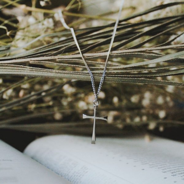 Silver cross pendant necklace suspended above an open Bible with soft dried florals and natural greenery, Christian faith jewelry, elegant silver cross necklace, inspirational religious pendant, devotional Christian accessory, spiritual gift for women and men, minimalist cross charm, Bible-themed jewelry photography, meaningful faith-based keepsake.