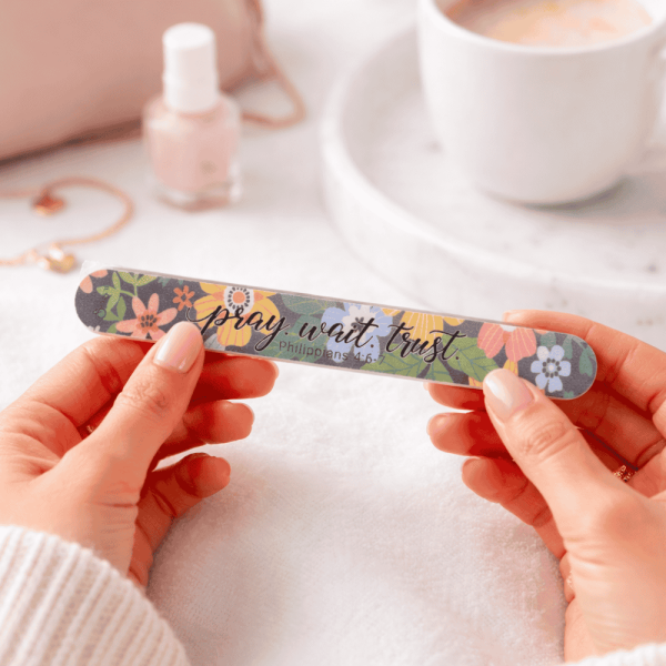 Hands holding floral Christian nail file with ‘Pray Wait Trust – Philippians 4:6’ message on soft aesthetic desk, faith-inspired gift accessory
