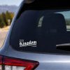 White vinyl decal featuring the phrase “Your Kingdom Come” in a bold Gothic-style font applied to the center of a silver laptop lid. The laptop sits on a clean wooden desk with a notebook, pen, potted plant, and coffee mug in a bright minimalist workspace with soft natural light.