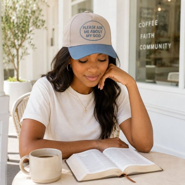 Woman wearing a beige and blue Christian baseball cap reading “Please Ask Me About My God” while enjoying coffee and reading her Bible at an outdoor café.