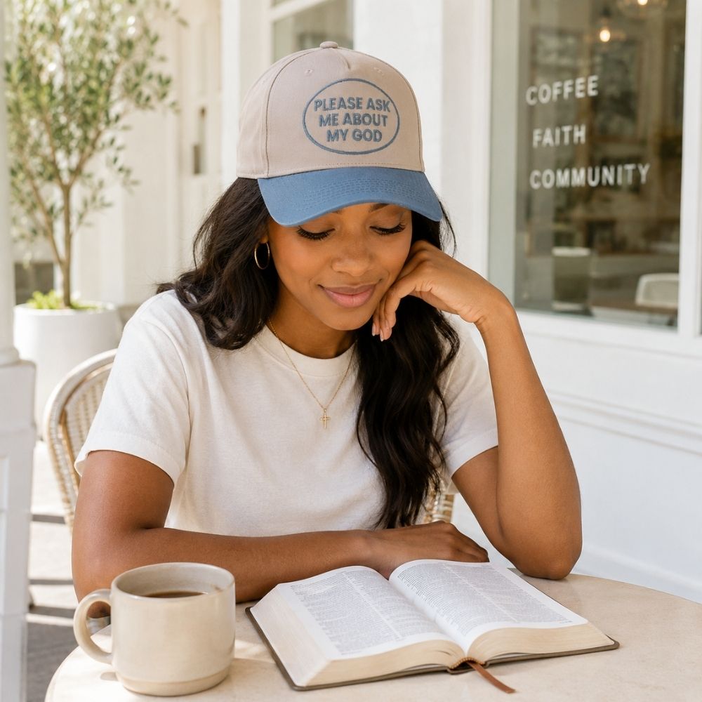 Woman wearing a beige and blue Christian baseball cap reading “Please Ask Me About My God” while enjoying coffee and reading her Bible at an outdoor café.
